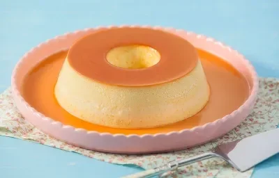 Foto de um pudim de leite moça dentro de prato rosa com bordas em cima de um pano colorido