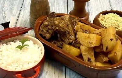 Foto em tons de marrom da receita de costela na pressão servida com batatas couradas em uma travessa com material que imita barro. Ao lado, uma panelinha vermelha com arroz branco e um recipiente com farofa