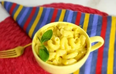 Foto da receita do mac and cheese na caneca servida em uma caneca amarela sobre um pano listrado colorido e ao lado um garfo dourado