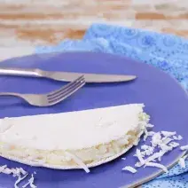 Fotografia em tons de azul em uma bancada de madeira clara, um pano azul, um prato azul redondo com uma tapioca de coco em cima dele.