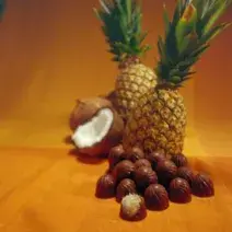 Fotografia em tons de marrom de uma bancada de marrom, sobre ela bombons, abacaxi e coco.