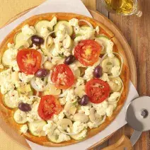 Fotografia em tons de amarelo em uma bancada de madeira com um pano amarelo, uma tábua redonda de madeira e a pizza de queijo, cogumelos, tomates e azeitonas em cima dela. Ao lado, um cortador de pizza.
