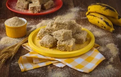 Imagem aproximada de uma mesa de madeira, com uma toalha quadriculada amarela e branca, um prato de madeira amarelo com a receita de palha italiana, algumas bananas inteiras e farinha láctea espalhada pela bancada ao redor