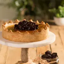 Fotografia em tons de marrom em uma bancada de madeira com um suporte de bolo branco com a torta manjar em cima. Ao lado, um potinho de vidro com ameixas secas pretas.
