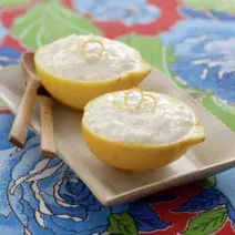Fotografia em tons de azul, amarelo e marrom de um paninho azul estampado com um prato beje, sobre ela duas metades de um  limão com o arroz doce, uma raspa da limão. Ao lado duas colheres de madeira.