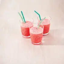 Fotografia em tons de rosa em uma bancada de madeira clara com três copos de vidro com o smoothie de melancia dentro deles e canudinhos verdes dentro dos copos.