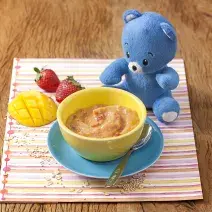 Fotografia em tons de amarelo em uma bancada de madeira e um paninho listrado colorido com um potinho amarelo com o creminho de manga com morango apoiado em um prato azul. Ao lado, um ursinho de pelúcia azul, dois morangos e uma fatia de manga.