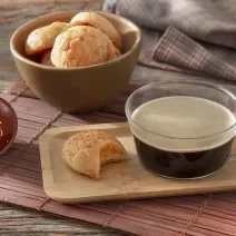 Foto da receita de pão de queijo 3 ingredientes servida em um bowl de cerâmica e à frente há uma xícara de café nescafé dolce gusto caffe matinal com um pão de queijo ao lado sobre uma tábua de madeira