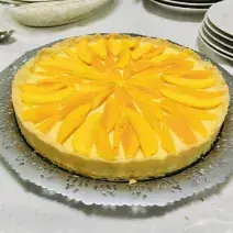 Fotografia em tons de amarelo em uma bancada de madeira de cor branca. Ao centro, uma mesa contendo a torta de manga. Ao fundo, alguns pratos brancos empilhados.