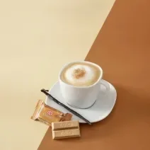 Foto da receita de cappuccino vanilla servida em uma xícara branca com um kit kat churros ao lado em fundo marrom claro e escuro