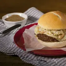 Fotografia em tons de azul e vermelho em uma bancada de madeira escura, um pano branco com listras azuis, um prato vermelho redondo raso e o hambúrguer recheado com queijo e mostarda em cima dele.