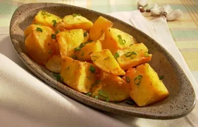 Fotografia em tons de amarelo em uma mesa com uma toalha quadriculada em amarelo, laranja e verde e um recipiente oval fundo com batatas cozidas e cebolinha-verde dentro dele.