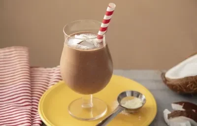 Fotografia em tons de marrom em uma bancada de madeira de cor cinza. Ao centro, um prato amarelo contendo uma taça com o smoothie. Ao fundo, há um pano branco listrado de vermelho e um coco partido ao meio.