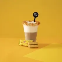 Foto em tons de amarelo da receita de latte salted caramel servida em um copo grande de vidro com um KIT KAT banana ao lado em fundo amarelo