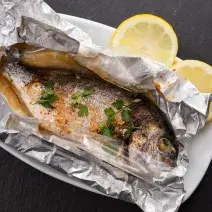 Fotografia em tons de cinza em uma bancada de madeira de cor preta. Ao centro, um prato branco contendo o peixe em um papel alumínio com duas rodelas de limão ao lado.