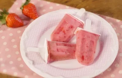 Fotografia em tons de rosa com um fundo de madeira e paninho rosa claro com bolinhas brancas, ao centro um prato branco pequeno e redondo com três sorvetinhos de morango. Ao fundo dois morangos inteiros decorando a imagem.