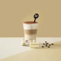 Foto da receita de Latte Tiramisú. Observa-se um fundo bege com o copo ao centro, dividido em 3 fases: embaixo, uma mais clara, a do meio, mais escura - de café, e a de cima, com creme. Por fim, chocolate polvilhado. À frente, um KITKAT de chocolate branc