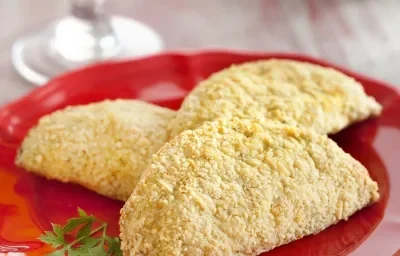 Fotografia em tons de vermelho em uma bancada de madeira com um pano branco e detalhes vermelhos, um prato vermelho redondo raso com três pasteis assados de frango. Ao fundo, uma taça vermelha com água.