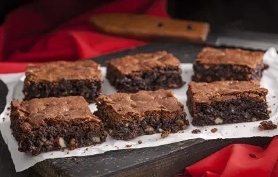 Foto de 6 pedaços iguais de brownie em cima de uma toalha branca e com uma faca de serra ao fundo da foto