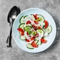 Fotografia vista de cima de uma salada de pepinos, tomates, cebola e grãos pretos por cima dentro de um prato fundo na cor azul. Ao lado, um garfo e uma colher, sobre uma mesa em tons de cimento.
