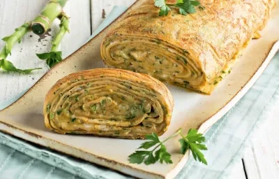 Fotografia em tons de verde em uma bancada de madeira clara com um recipiente retangular bem comprido e grande com a omelete em forma de rocambole.