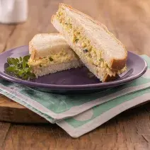 Foto em tons de bege da receita de pãozinho com ovo mexido servida em um prato de porcelana roxo, sendo duas metades do sanduíche, uma por cima da outra. O prato se encontra sobre uma tábua de madeira com um paninho verde claro entre eles.