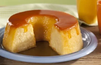 Fotografia em tons de bege e verde de uma bancada de madeira com um prato lilás com o pudim cortado, ao fundo um paninho verde quadriculado verde e branco e um copo amarelo.