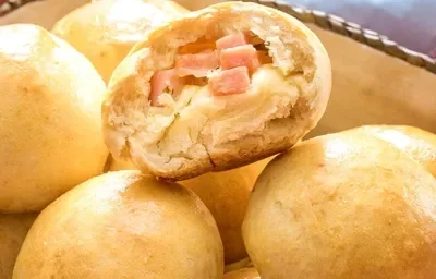 Fotografia em tons de amarelo em uma bancada de madeira vermelha um pano azul, uma cesta de vime com pães recheados com queijo.