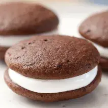 Fotografia em tons de preto em uma bancada de madeira clara, um prato redondo branco raso com três sanduicinhos de chocolate recheados com marshmallow.