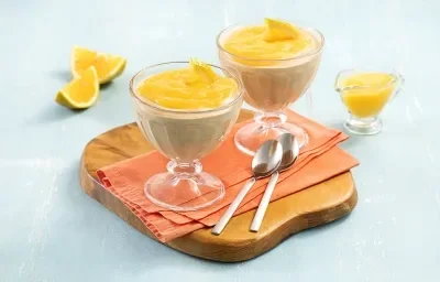 Foto da receita de Mousse de Cappuccino com Calda de Laranja. Oberva-se duas taças transparentes com a mousse e a calda laranja por cima em cima de um guardanapo laranja e uma tábua redonda de madeira. Como decoração, pedaços de laranja em um fundo azul.