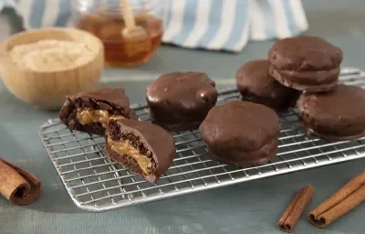 Foto da receita de pão de mel servida em diversas porções usando uma grade como base acima de uma mesa azul clara com paus de canela para decoração, além de uma cumbuca com farinha láctea e um pote de vidro com mel