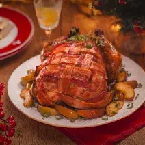 Fotografia em tons de vermelho em uma bancada de madeira escura, um pano vermelho, um prato branco redondo com o chester coberto com bacon em cima dele. Ao lado, copos, pratos e decoração e enfeites natalinos.