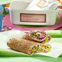 Fotografia em tons de verde em uma mesa verde com um pano verde, um papel manteiga com dois wraps recheados com salada e cebola. Ao fundo, uma marmita branca com detalhes rosa.