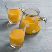 Fotografia em tons de azul e amarelo com uma jarra de vidro com bolinhas brancas e dois copos contendo suco de frutas, tudo sobre bancada em tons de azul.