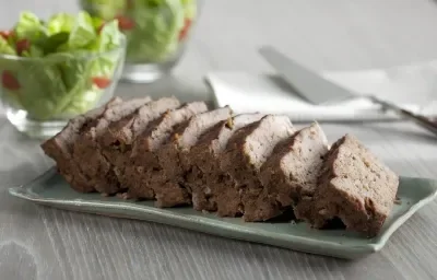 Fotografia em tons de marrom e cinza, com bolo de carne assada sobre peça de porcelana cinza e no entorno guardanapo branco com espátula por cima e dois potinhos com folhas verdes, tudo sobre bancada em tons de cinza.