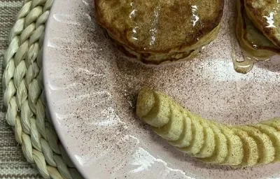 Foto da receita de Panquequinha de Aveia. Observa-se um prato com panquequinhas com mel e canela e do lado de baixo do prato, uma banana em rodelas para formar um sorriso.