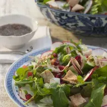 Fotografia em tons de verde em uma bancada de madeira com um prato branco com detalhes em azul e a salada com agrião, rabanete, alface dentro. Ao fundo, um potinho com molhinho.