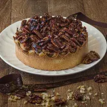 Foto da receita de Torta de Caramelo Salgado e Castanhas Caramelizadas, servida num prato redondo branco, sobre uma bancada de madeira, decorada com uma fita de páscoa e castanhas picadas