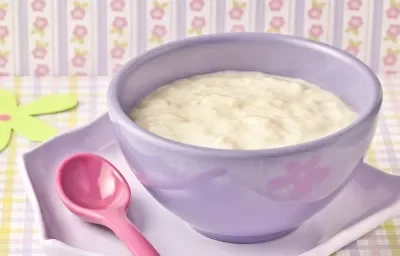 Fotografia em tons de lilás, com pote lilás ao centro com creme de banana, sobre prato lilás em forma de pentágono e colher rosa, sobre toalha quadriculada em branco, amarelo e lilás, e papel de parede colorido e com motivos florais. Fotografia em tons de lilás, com pote lilás ao centro com creme de banana, sobre prato lilás em forma de pentágono e colher rosa, sobre toalha quadriculada em branco, amarelo e lilás, e papel de parede colorido e com motivos florais.