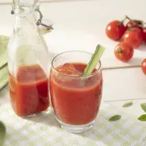 Fotografia em tons de vermelho em uma bancada de madeira clara, um pano verde liso e outro xadrez, um copo de vidro com o suco de tomate e uma jarra com mais suco ao centro da foto. Ao lado, tomates pequenos para decorar a mesa.