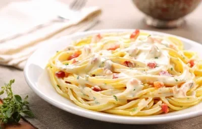 Fotografia de um prato de macarrão cozido, coberto com molho branco cremoso e bacon crocante.