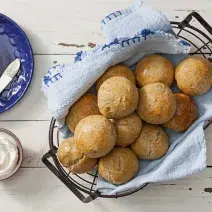 Fotografia em tons de branco e azul de uma bancada branca com uma cesta, dentro dela um paninho azul e pâozinhos. Ao lado um potinho com requeijão e um prato pequeno azul com um pão e uma faca.