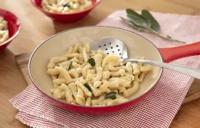 Foto da receita de Spatzle. Observa-se uma frigideira grande sobre um pano de prato quadriculado vermelho. A massa está dentro da frigideira com folhas de manjericão.