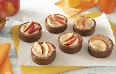 Foto da receita de Tortinha de Maçã. Observa-se 6 tortinhas decoradas com rodelas de maçã no topo sobre um papel-manteiga e uma tábua amarela. Foto da receita de Tortinha de Maçã. Observa-se 6 tortinhas decoradas com rodelas de maçã no topo sobre um papel-manteiga e uma tábua amarela.