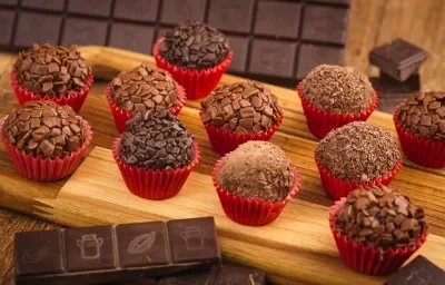 Fotografia em tons de vermelho em uma bancada de madeira com vários brigadeiros colocados em forminhas vermelhas em cima de uma tábua de madeira clara. Ao fundo, uma barra de chocolate meio amargo e com alguns pedaços de chocolate espalhados.