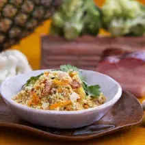 Foto da receita de farofa de casca de abacaxi com talos, servida em um prato fundo, sobre outro prato marrom, numa bancada alaranjada onde há um abacaxi grande ao fundo