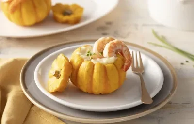 Fotografia em tons de laranja de uma bancada, ao centro dois pratinhos pequenos e dentro dele uma mini abóbora recheada e decorada com dois camarões. Ao lado um garfo, paninho amarelo e ao fundo outra mini abóbora. Fotografia em tons de laranja de uma bancada, ao centro dois pratinhos pequenos e dentro dele uma mini abóbora recheada e decorada com dois camarões. Ao lado um garfo, paninho amarelo e ao fundo outra mini abóbora.