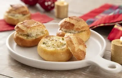 Foto da receita de Pão de Alho na Air Fryer. Observa-se três pãezinhos italianos em um recipiente refratário branco de porcelana, sendo que o da frente está aberto expondo o recheio de queijo.