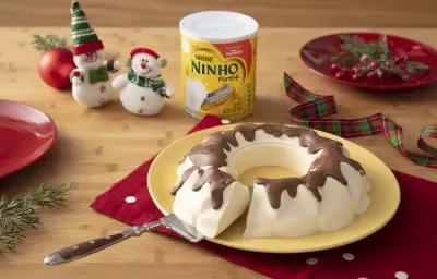 Fotografia em tons de vermelho e branco de uma bancada de madeira, um paninho vermelho e um prato amarelo com o bolo de sorvete com calda de chocolate em cima. Ao fundo decoração de natal, com mini bonecos de neve e uma lata de NINHO.