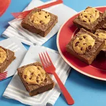 Fotografia em tons de azul e vermelho de uma bancada azul com paninhos azul claro com listras brancas e sobre eles pedaços de brownie com cookies e garfos vermelhos. Ao lado um prato vermelho e sobre eles pedaços de brownie com cookies.